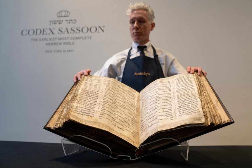 A man in an apron holds a large, weathered old book up for display.