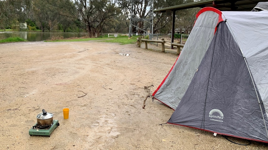 A tent on a gravel campground with a billy and gas stove brewing tea beside it. In the background the Wodonga creek. 