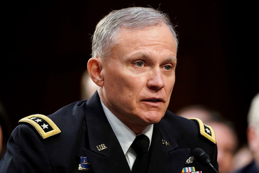 A US Lieutenant General in dress uniform speaks into a microphone while seated at a table