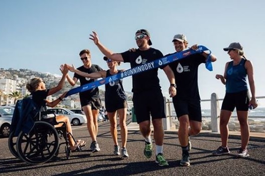 Mina Guli sits in a wheelchair on the left of frame as five of her team members cross the finish line of marathon near beach.