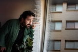 A man with dark curly hair peeks through the blinds of his apartment window.