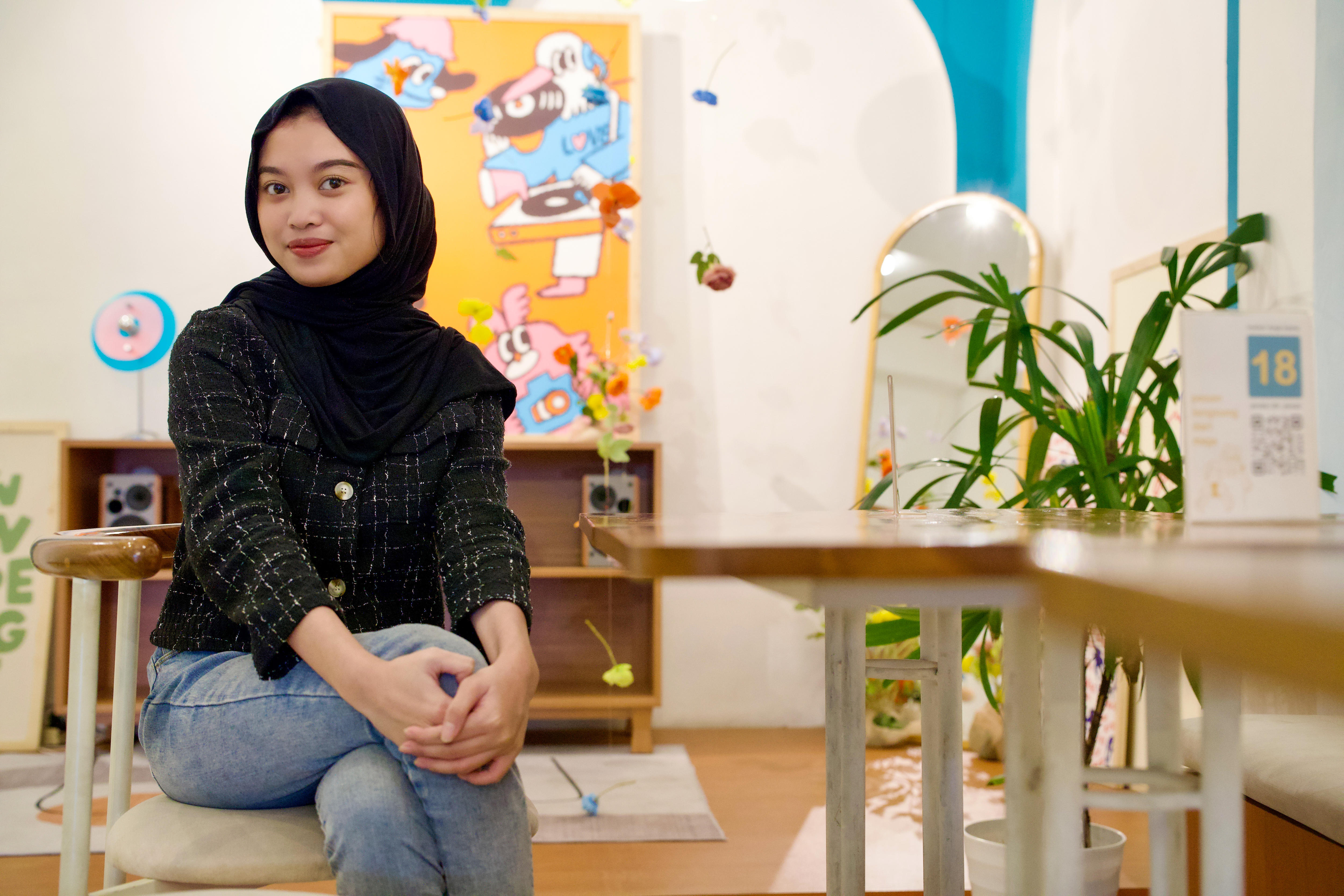 A woman wearing a hijab poses while seated on a chair in a bright room.