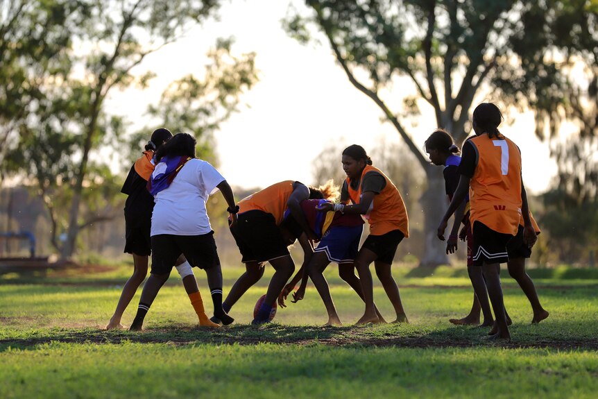 A group of young Aboriginal women wearing football jumpers compete for a ball on the ground in the late afternoon sun