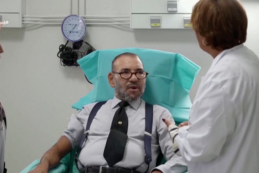 King Mohammed VI looks at a doctor as they prepare his arm to donate blood.