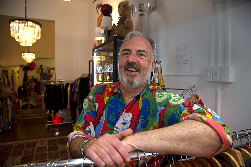 Man with bright blue eyes and a colourful shift smiles with one arm on a clothes rack.