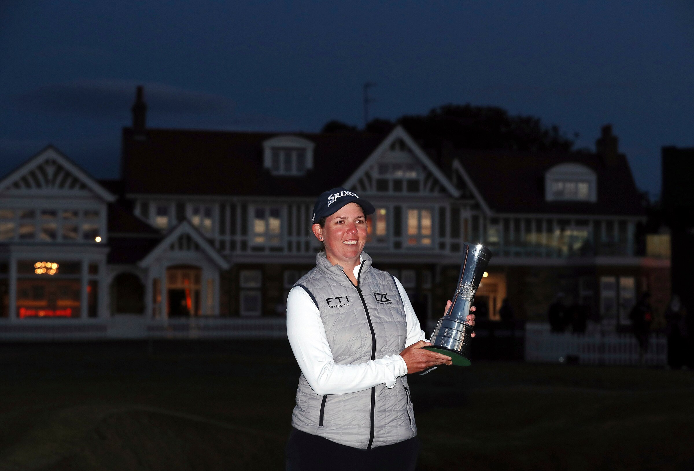 A golfer smiles as she stands in the light holding a major trophy, with a clubhouse in near-darkness behind her. 
