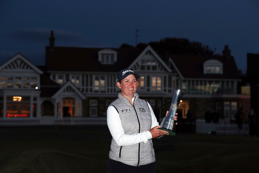 A golfer smiles as she stands in the light holding a major trophy, with a clubhouse in near-darkness behind her. 
