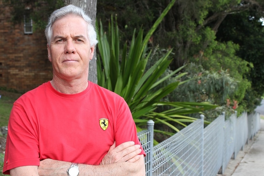 A man with grey hair and a red t-shirt, standing with his arms folded.