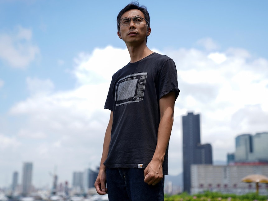 Un bărbat care poartă ochelari stând afară pe fundalul orizontului Hong Kong-ului 