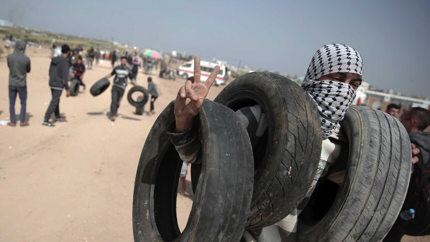 A man covering his face with a Palestinian scarf is seen carrying tires and gesturing the victory hand sign.