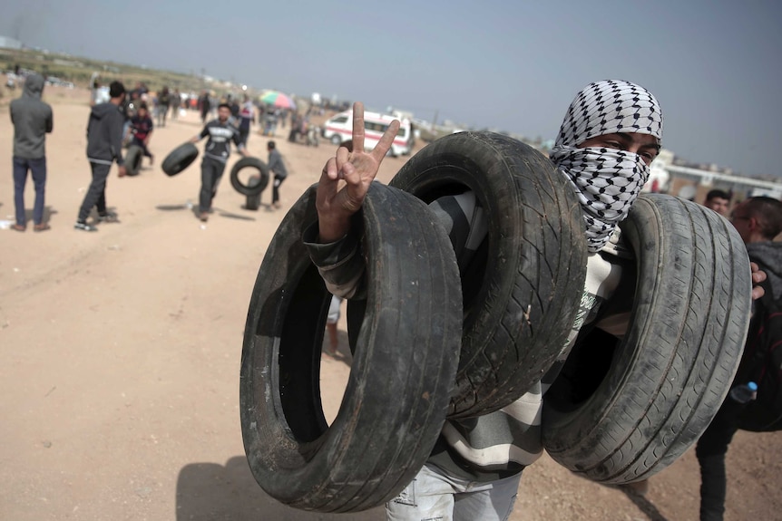 A man covering his face with a Palestinian scarf is seen carrying tires and gesturing the victory hand sign.