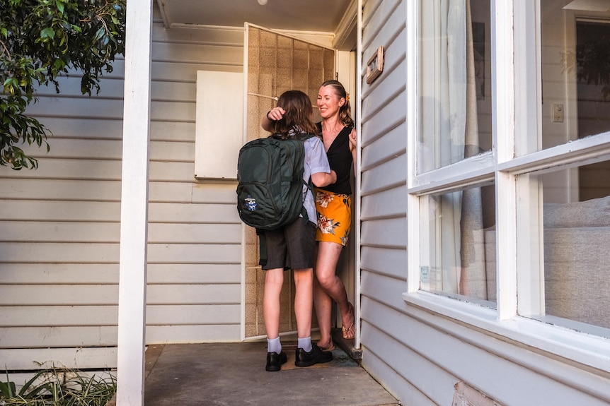 A mother says goodbye to a school-aged child at the front door of a house.