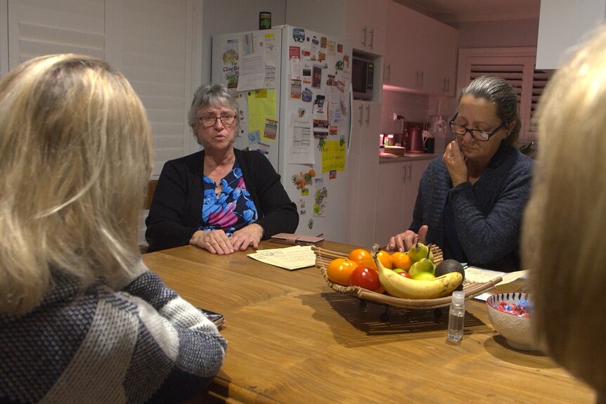 Four women sit around a dining room table, one is talking. Behind them is a kitchen.