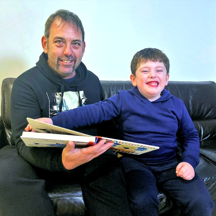 Andrew Bird and his son Isaac sit together on the couch with a picture book while smiling at the camera