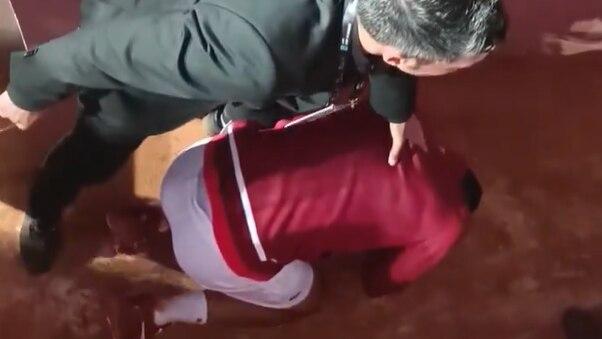 An athlete in a red shirt kneels low on the ground clutching his head as a man in a suit stands over him.