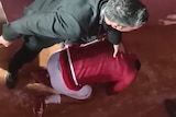An athlete in a red shirt kneels low on the ground clutching his head as a man in a suit stands over him.