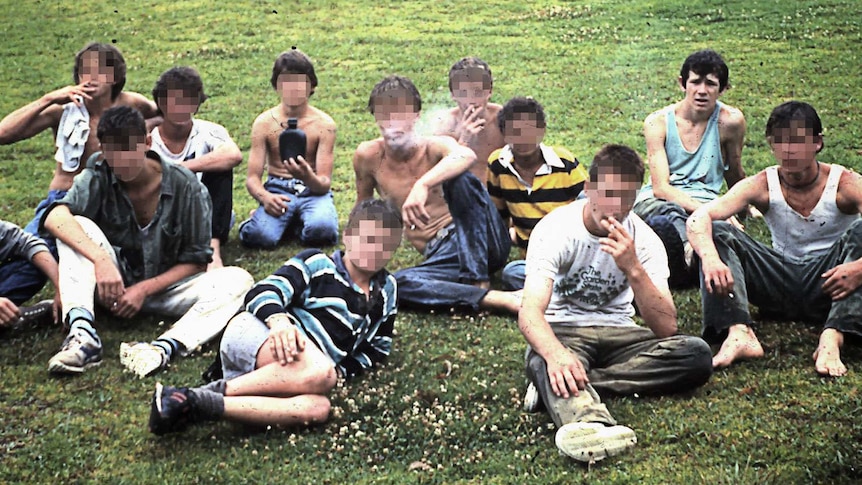 A group of teenage boys sit in a field smoking cigarettes.