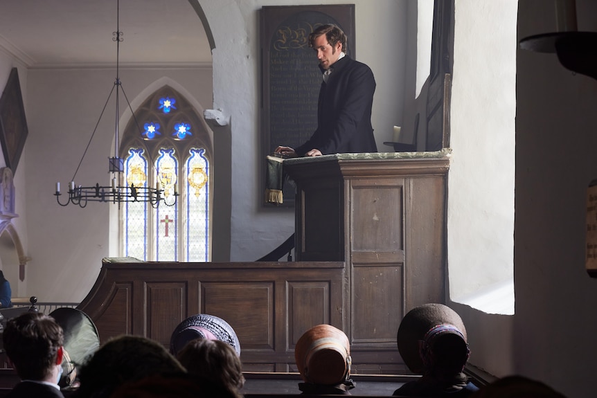 White man with dark hair wears a black coat and stands at a wooden podium inside a church before a congregation.