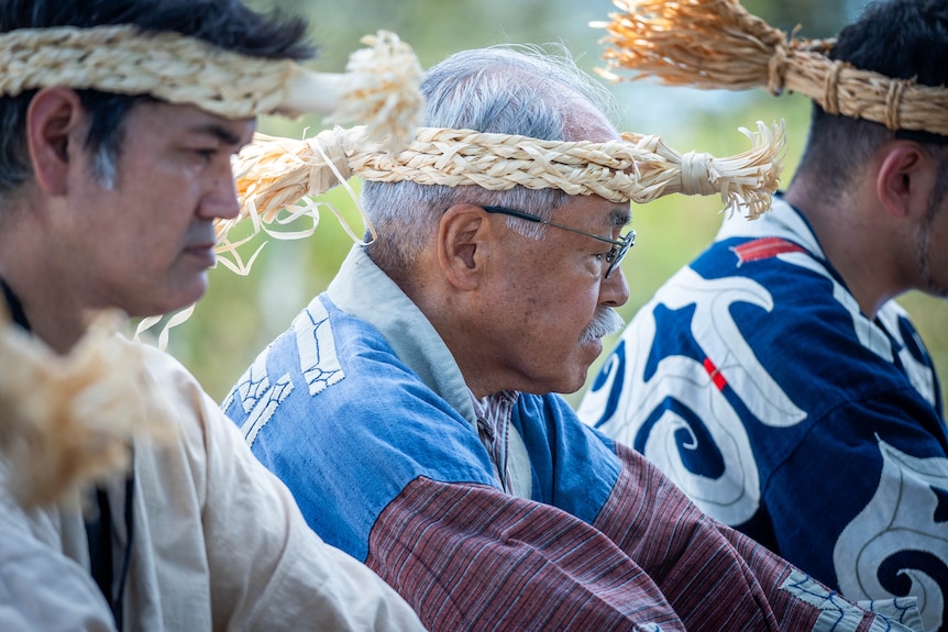 Three men in woven headdresses sit in a row 