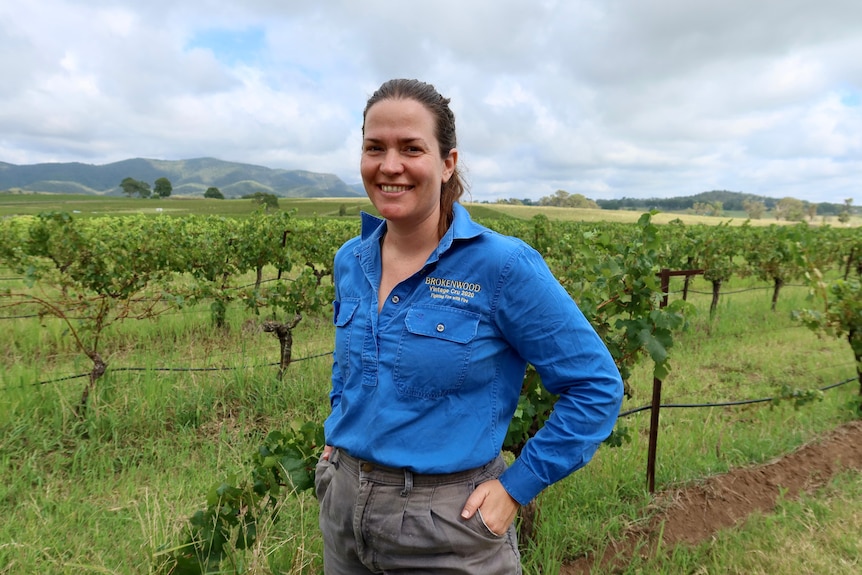 A young woman in a long-sleeved blue shirt smiles as she stands near vineyards.