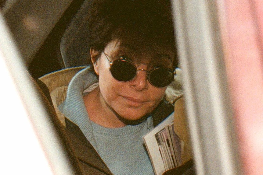 A woman with cropped hair and dark shades sits in the back of a police car. The photo is taken through the door