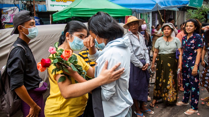 A young woman holding flowers cries as another woman comforts her and others look on.