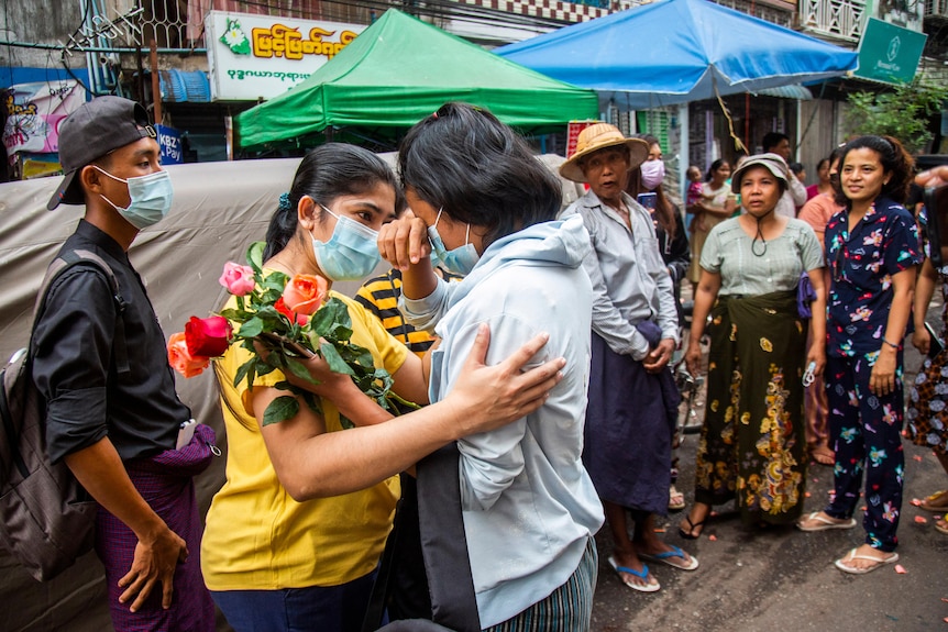 A young woman holding flowers cries as another woman comforts her and others look on.