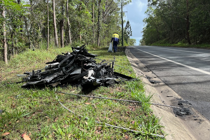 A mangled pile of metal – the result of a car crash – on the side of a country road. A council worker collects debris behind it.