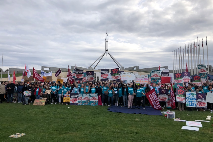 a crowd of protesters holding signs gather on the lawn outside Parliament House.