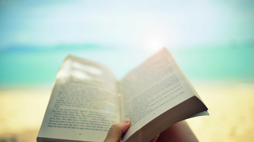 A hand holds a book open, with an out of focus view of the beach in the background.