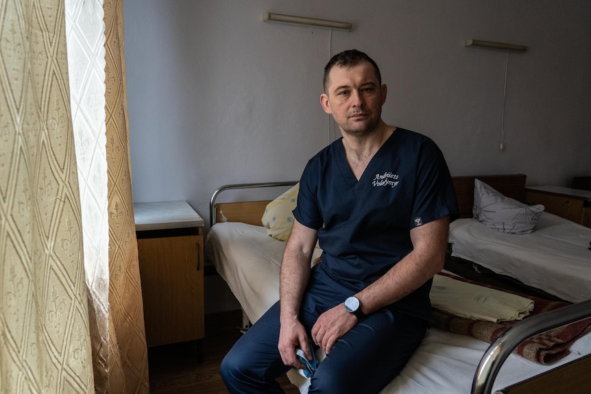 A doctor in blue scrubs sits on a hospital bed.