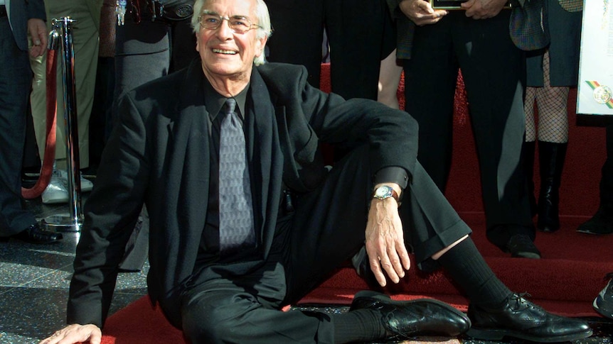 Actor Martin Landau sits on his star on the Hollywood Walk of Fame posing for photographs as with a wall of people behind him.