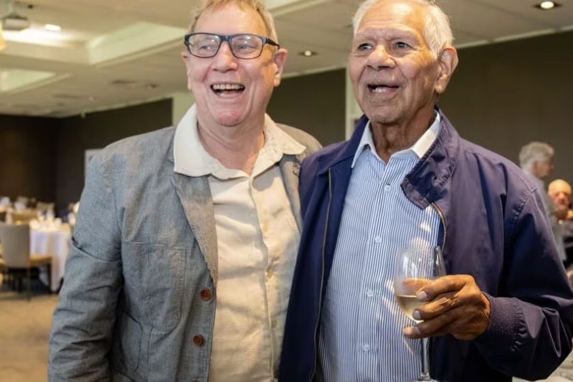 Two older men stand together, laughing.