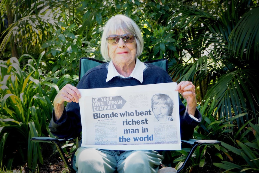 A smiling older woman sits in a verdant garden, holding a newspaper article headlined 'Blond who beat Richest Man in the World'.
