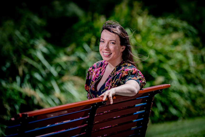 A smiling woman with a floral dress in the sunshine. She is sitting on a park bench