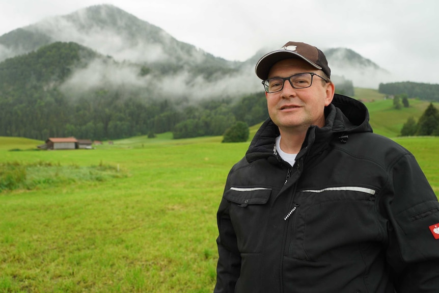 A man wearing glasses and a baseball cap stands in front of a green landscape with fields, a barn and hills in the background