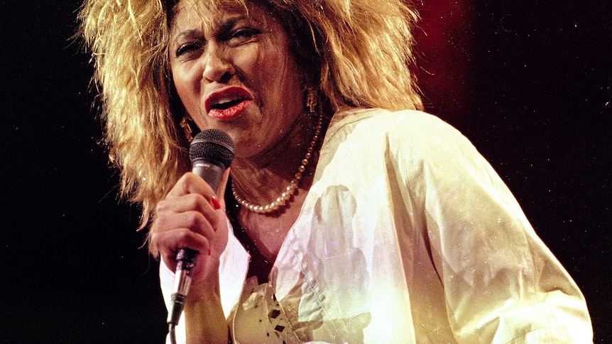 Tina Turner sings into a microphone, wearing a white jacket and with large hair bleached blonde.