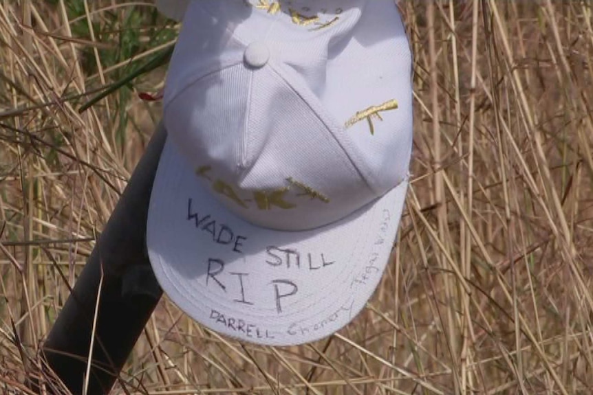 A white hat cap on a pole in front of brown grass. It reads "Wade Still RIP".