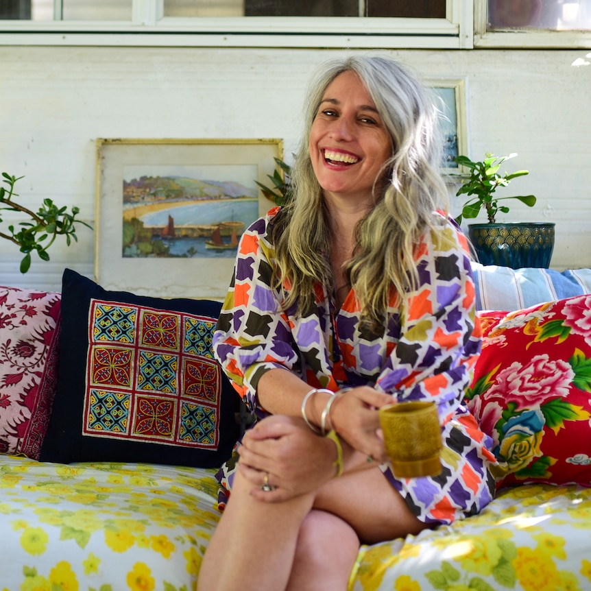 A woman with long, silver hair sitting on a couch with colourful cushions.