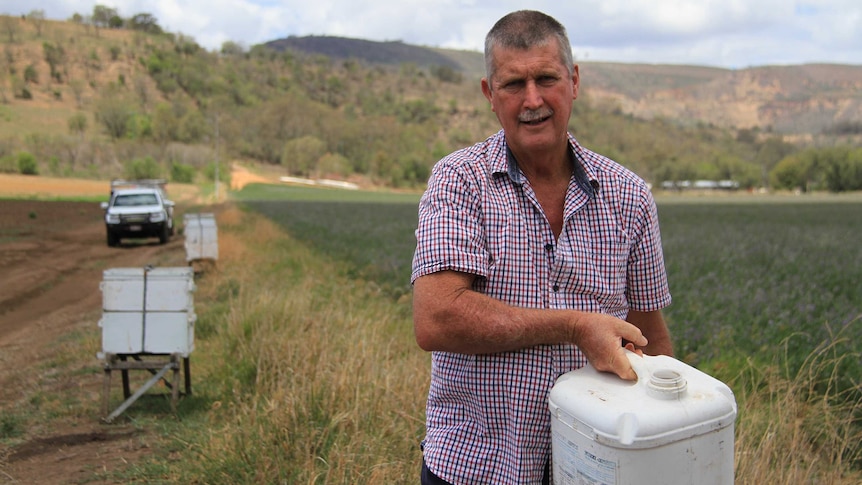 Phil Dunlop stands in front of beehives and small crops, holding a drum of water.
