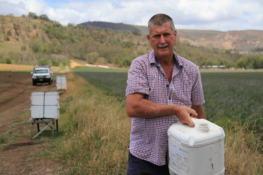 Phil Dunlop stands in front of beehives and small crops, holding a drum of water.