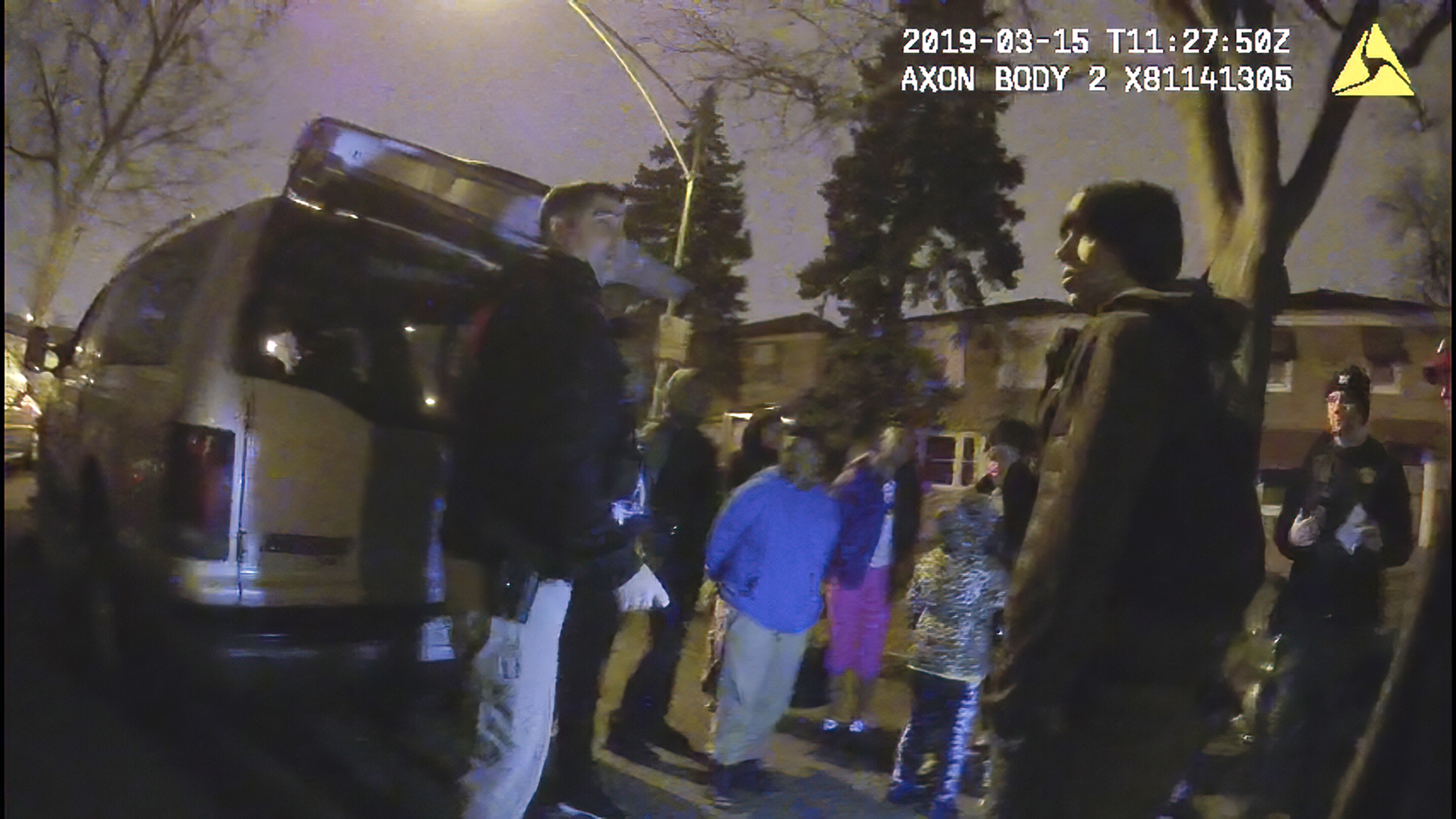 Young boy wearing blue handcuffed, surrounded by police and family.