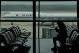 A passenger checks her phone in an airport terminal, with an Air China plane on the tarmac outside the terminal window.