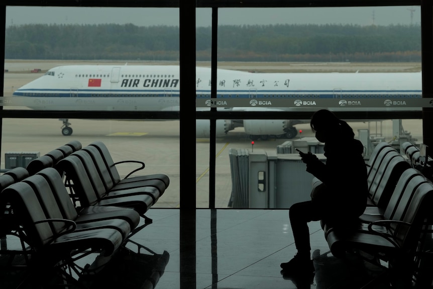 一名乘客在机场候机楼查看手机，候机楼窗外的停机坪上停着一架中国国际航空公司的飞机。