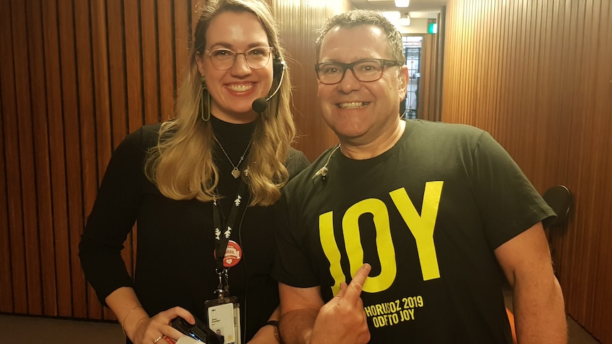 Lizzy Greenhalgh and Damien Beaumont post for a photo. Lizzy has a headset on and Damien points to the word 'JOY' on his tshirt.