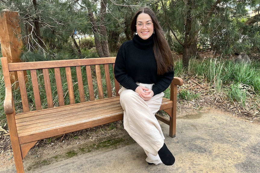 Young woman in black high neck jumper, wears glasses, smiling, long brown hair, sitting on park bench with trees behind her