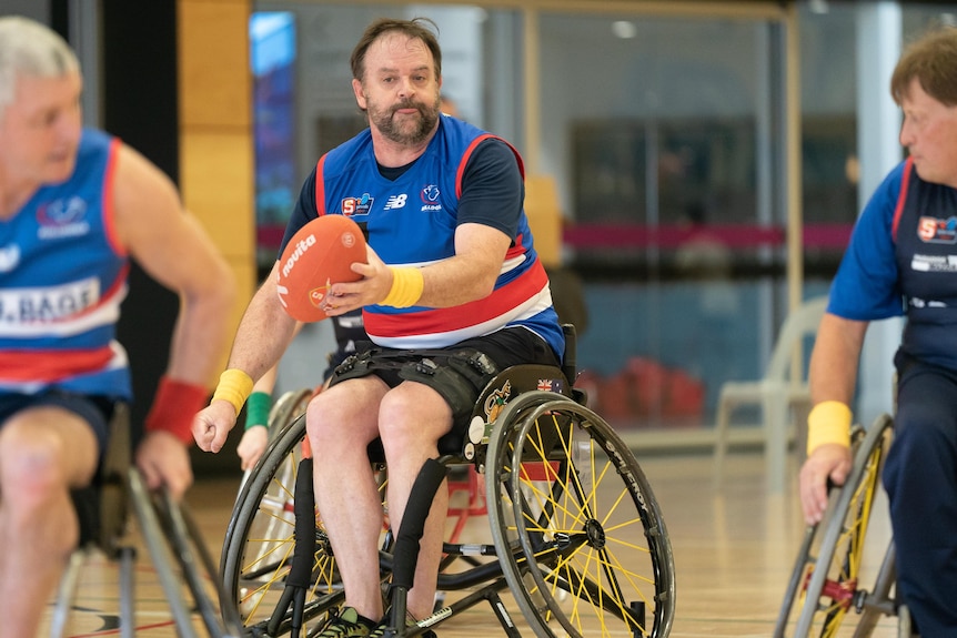 Un homme en fauteuil roulant se prépare à jouer au handball avec un coéquipier lors d'un match de football en fauteuil roulant alors qu'un adversaire regarde