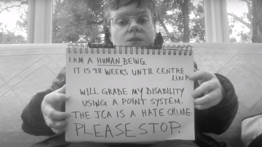 Black and white photo of Sophie holding a note book with a statement that includes "THE JCA IS A HATE CRIME, PLEASE STOP".