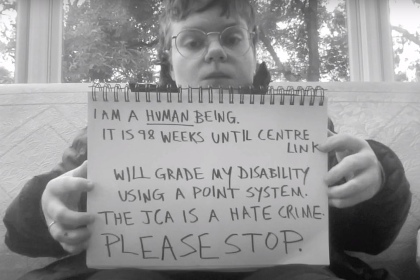 Black and white photo of Sophie holding a note book with a statement that includes "THE JCA IS A HATE CRIME, PLEASE STOP".