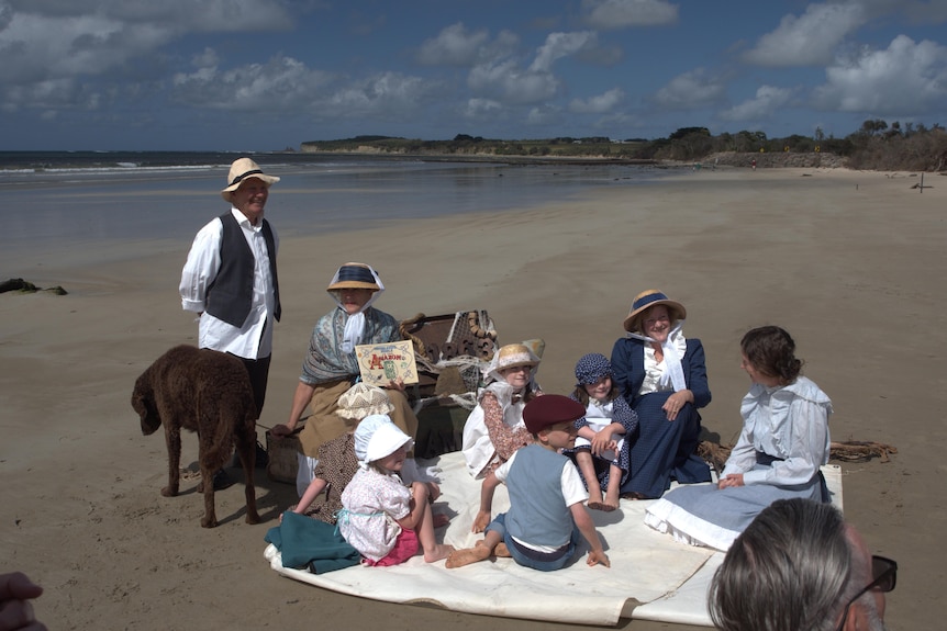 karyn is holdiong amazon book with adults and children around her all in period clothes, they are on the beach 